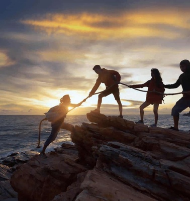 Four people holding hands and helping each other on a rocky shore during sunset.
