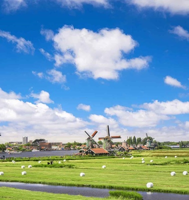 A scenic view of traditional Dutch windmills beside a river in a lush green field under a cloudy blue sky.