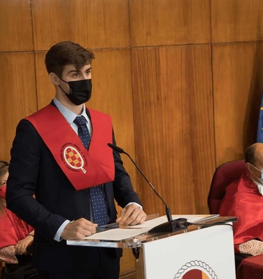 A young man in a red sash and black mask giving a speech at a podium, with seated individuals wearing red robes and masks in the background.