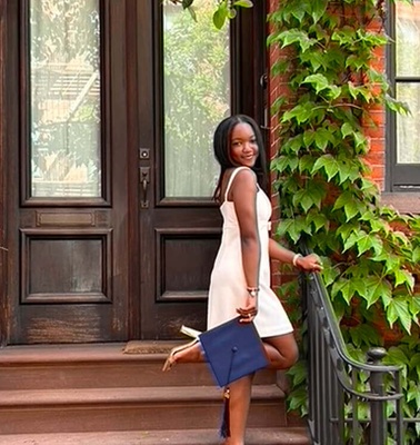 A young woman stands elegantly on the stairs of a charming brick building covered in green ivy.