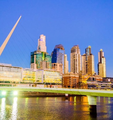 A modern bridge illuminated at dusk with a city skyline in the background.