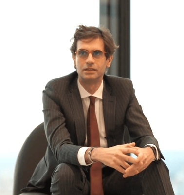 A man in a suit sits thoughtfully in a modern office environment, overlooking a cityscape through large windows.