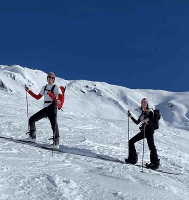 Two people cross-country skiing on a snowy mountain slope under a clear blue sky.