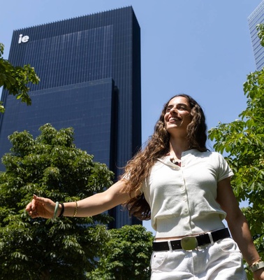 A smiling woman walking among green trees with a modern building in the background.
