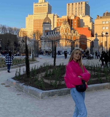 A woman in a pink jacket posing in a city park during the day, with city buildings in the background.