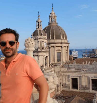 A man in sunglasses stands in front of a historic cathedral with a view of the harbor in the background.