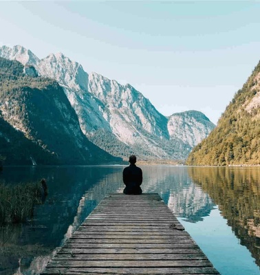 A person sitting at the end of a wooden dock overlooking a calm lake surrounded by mountains.