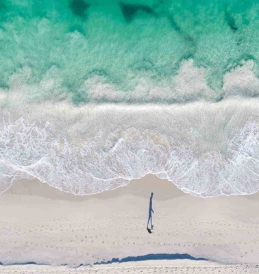 Aerial view of a single person walking on a sandy beach with turquoise water and white surf.