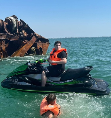 A person in a life jacket is riding a jet ski near a large rusted shipwreck in the ocean.