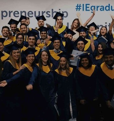 Group of graduates celebrating in blue and yellow regalia at a graduation ceremony, with a banner reading #IEgraduation in the background.