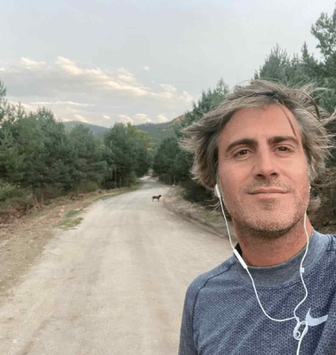 A man with gray hair taking a selfie on a rural road with trees and a distant dog.