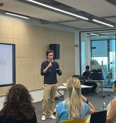 A speaker presents in front of an audience with a large screen showing illustrations.
