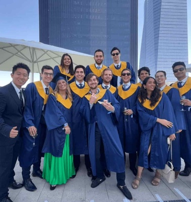 A group of graduates in blue robes celebrating outdoors with skyscrapers in the background.