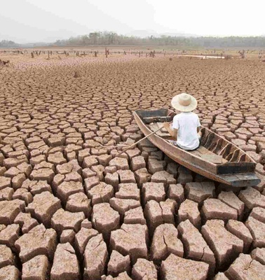 A person sits in a wooden boat stranded on a cracked dry lake bed.