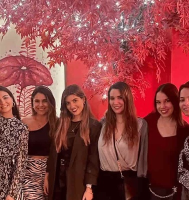 A group of seven women posing together in a room decorated with red foliage and a whimsical umbrella.