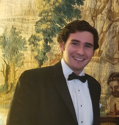 A young man in a tuxedo smiling in front of a decorative tapestry.