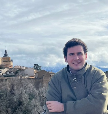 A man smiling in the foreground with a historic cityscape including a cathedral in the background under a cloudy sky.