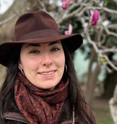 A woman wearing a brown hat and scarf smiles in front of a tree with pink flowers.
