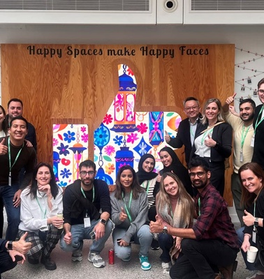 A group of people posing with thumbs up in front of a colorful mural that reads 'Happy Spaces make Happy Faces'.