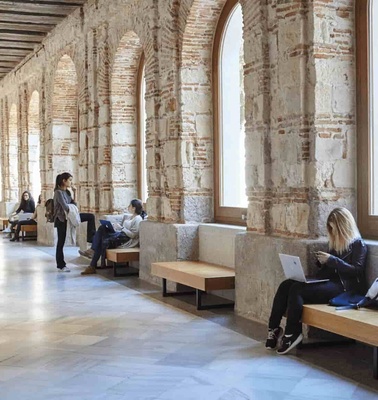 People sitting and standing in a historical hall with arched windows and a corridor.