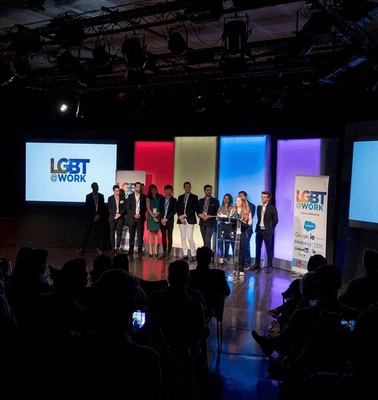 A group of people standing on a stage at an event, with colorful 'LGBT at Work' banners displayed in the background.
