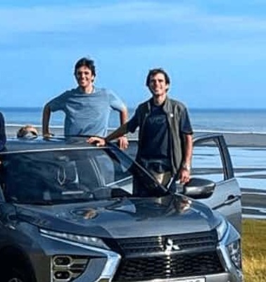 Four men standing in front of an SUV on a grassy coastal area with a wide view of the sea and sky in the background.