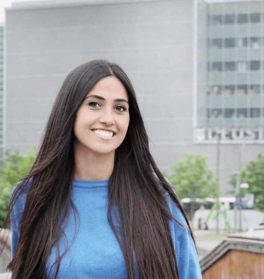A young woman with long dark hair smiling in front of a building with the Boston logo.