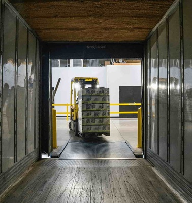 A pallet of goods secured with yellow straps, viewed inside the back of an open semi-truck, with a forklift nearby in a warehouse setting.