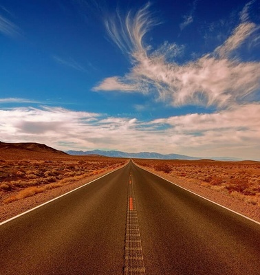 A long, straight road stretches into the horizon under a bright blue sky with wispy clouds.