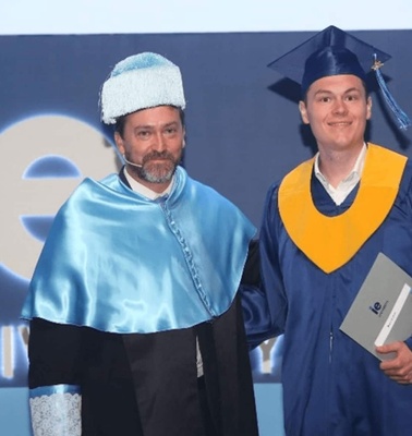 A graduation ceremony photo showing a male graduate in cap and gown holding a diploma standing next to a male academic in formal regalia.