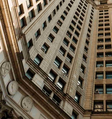 Looking up at a towering beige skyscraper flanked by ornate classical architecture under a clear sky.