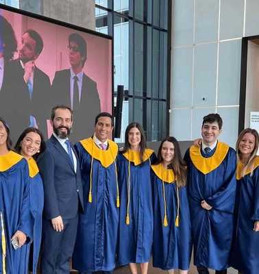 A group of eight graduates in blue and yellow gowns smiling with a professor in a suit, standing in front of a screen displaying an event.