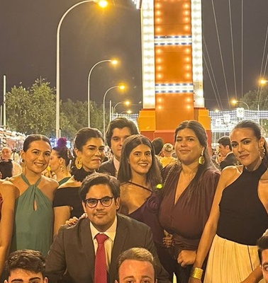 A group of people dressed in formal attire posing for a photo at a night event with bright, decorative lights in the background.