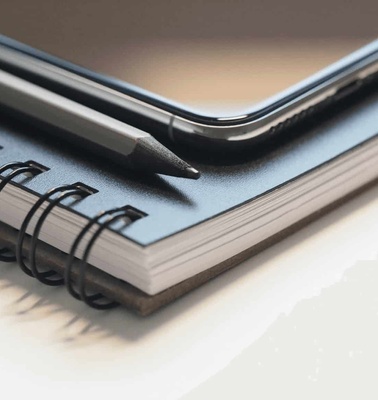 A close-up image of a smartphone laying on a spiral notebook with a pencil on top.