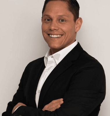 A smiling man in a black suit and white shirt posing against a light grey background.