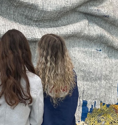Two women observe a textured artwork made of various materials.