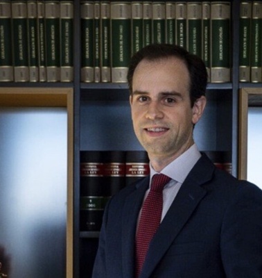 A man in a suit standing in a library with books and an open book on the table.