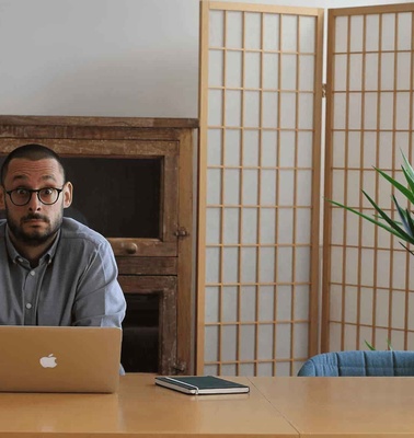 A man in glasses sitting at a desk with a laptop in a room with a wooden panel, a plant, and a Japanese-style screen.