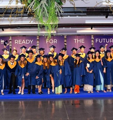 A group of graduates wearing blue robes and yellow stoles gather for a photo at a graduation ceremony.
