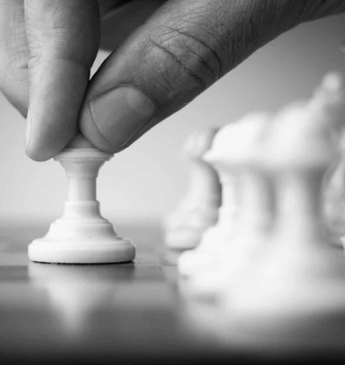 A black and white photo of a hand moving a pawn on a chessboard, with other chess pieces blurred in the background.