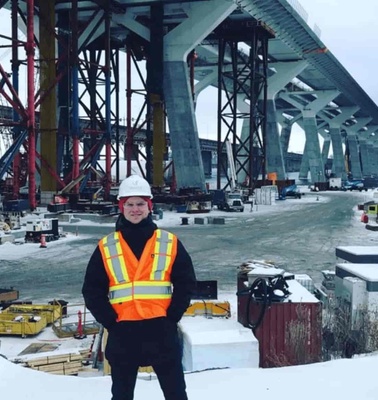 A person in a high-visibility vest standing in front of a large bridge under construction during winter.