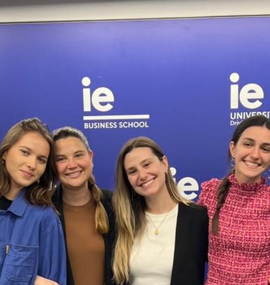 A group of six young adults smiling in front of a banner with logos of IE University and Business School.