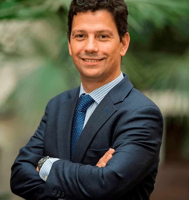 A professional man in a suit smiling with his arms crossed in front of a green plant background.