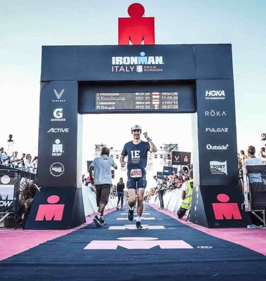 A runner crossing the finish line at an Ironman event in Italy, with cheering spectators on both sides.
