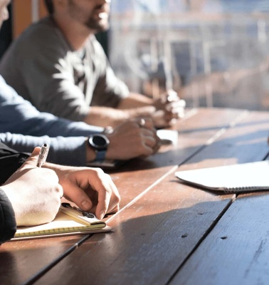 A group of people engaged in a meeting at a wooden table, focusing intently with notebooks and pens.