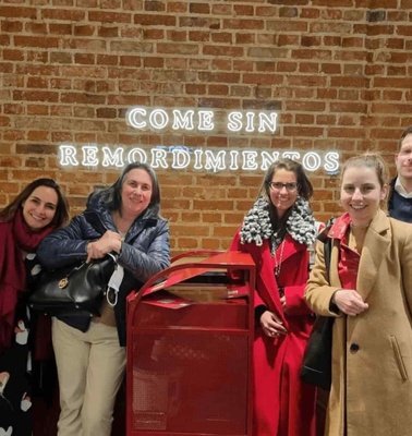 Five people posing happily in front of a brick wall with a neon sign that reads 'COME SIN REMORDIMIENTOS'