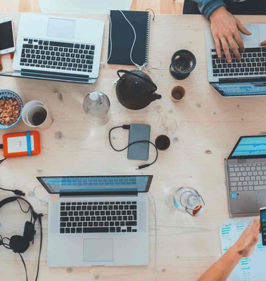 Aerial view of a busy workspace with multiple people typing on laptops and various objects like notebooks, mobile phones, and earphones scattered around.