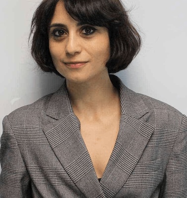 A woman with short dark hair wearing a gray blazer stands against a plain background.