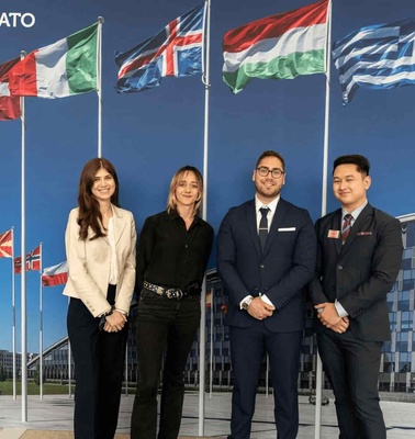 Four people standing in front of multiple national flags outside a building, under a sign saying '#WEARENATO'.