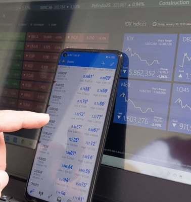 A person comparing financial data on a smartphone and a laptop screen, both displaying stock market charts and numbers.